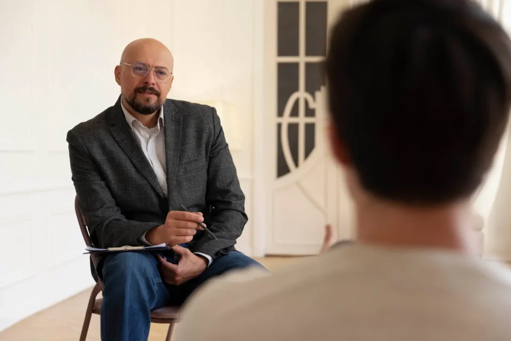 Bald man with glasses and a beard, dressed in a grey blazer, holding a pen and clipboard, sitting across from another person in a bright healthcare facility.