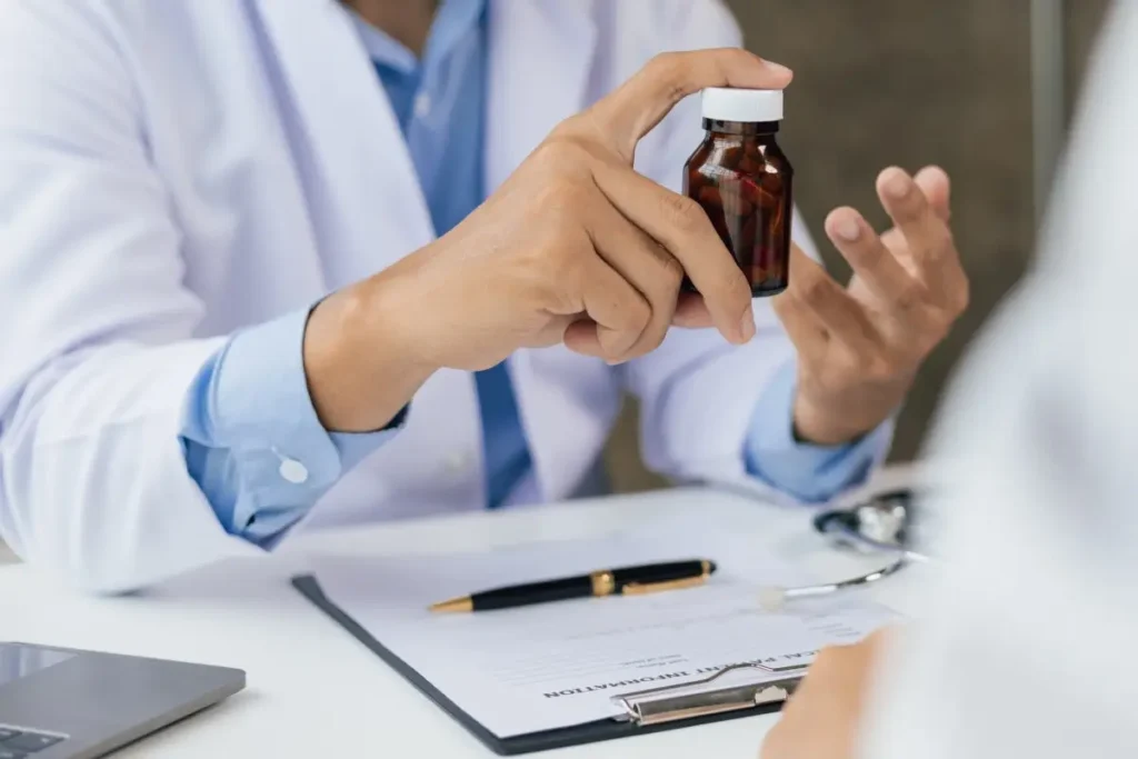 A doctor in a white coat holding a medicine bottle and explaining it to a patient, with a stethoscope and clinical documents on the desk in the foreground.