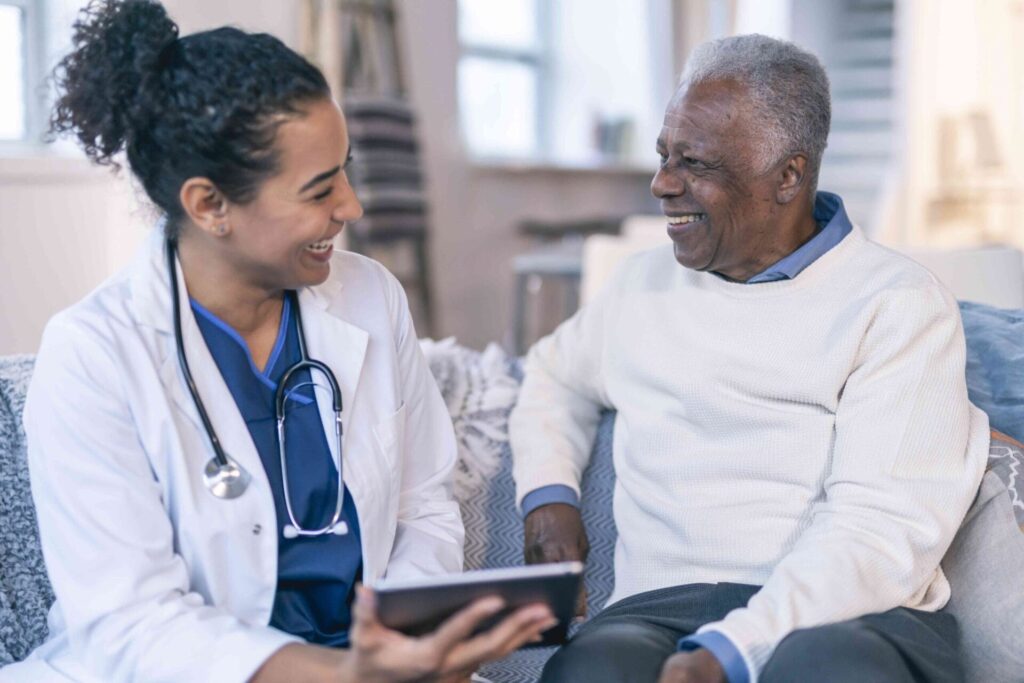 A female doctor with a stethoscope around her neck, smiling and showing a tablet to an elderly male patient, who is also smiling, as they sit in a bright living room for an Advanced Primary Care consultation.