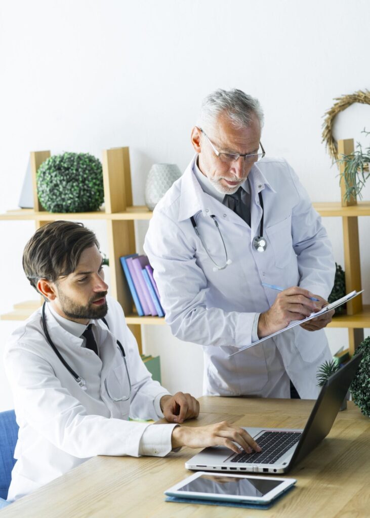 Two male doctors, one older with gray hair and the other younger with a beard, are focused on a digital tablet and laptop in a clinical medical office.
