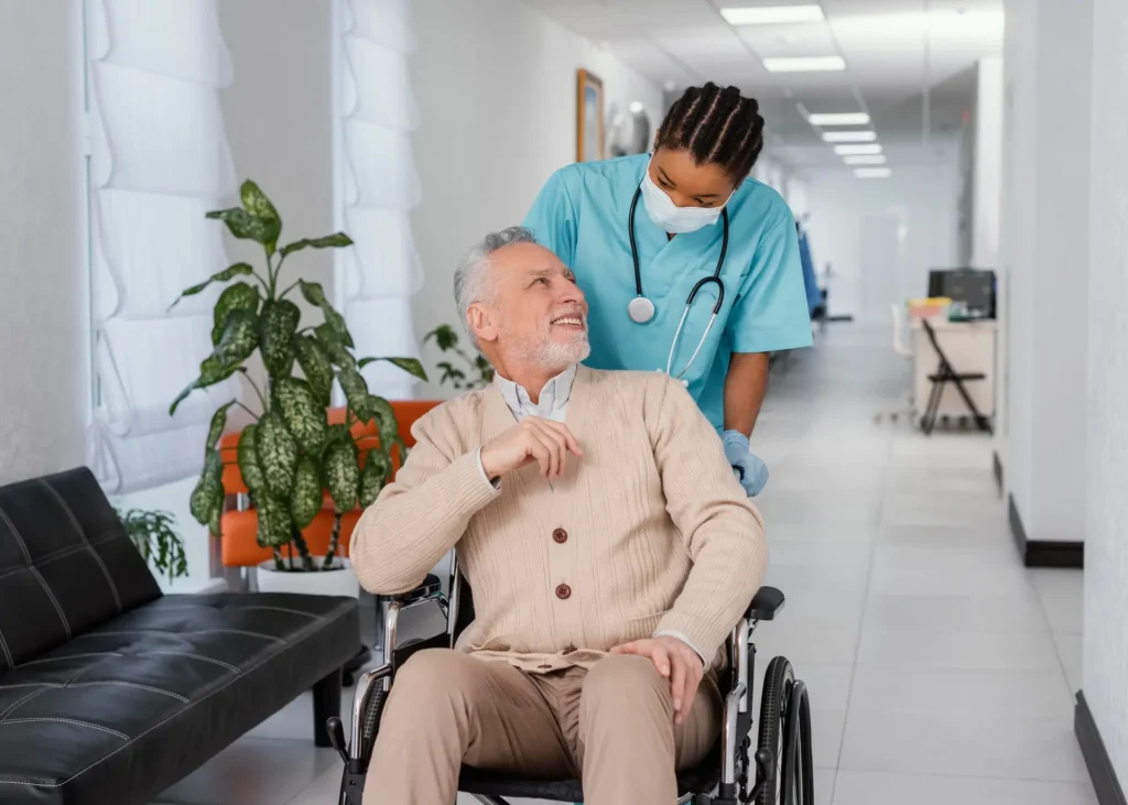 A healthcare professional is gently pushing an elderly man in a wheelchair down a bright hallway, as he looks up smiling, interacting joyfully with his primary care provider.