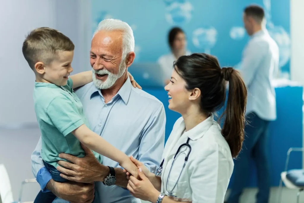 A joyful scene in a primary care clinic where an elderly man holds a young boy, laughing together, while a female doctor smiles and converses with them, all against a blurred background of medical staff.