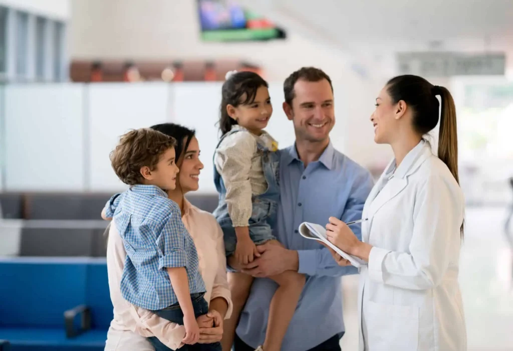 A happy family of four, including two young children and their parents, interacts with a female doctor holding a clipboard in a bright primary care clinic lobby.
