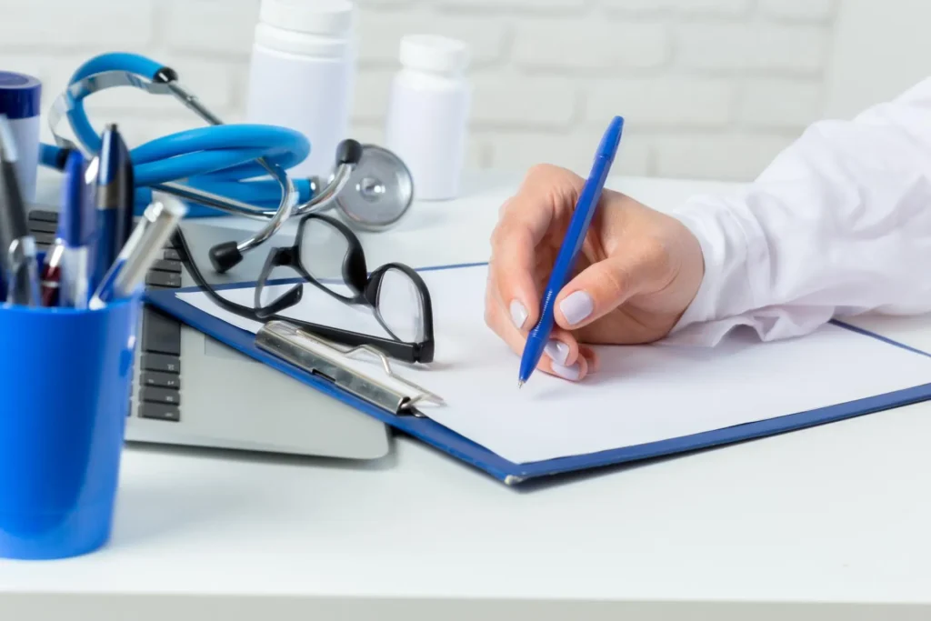 A healthcare professional writes on a clipboard in an Advanced Primary Care office, surrounded by medical equipment including a stethoscope, eyeglasses, and medication bottles.