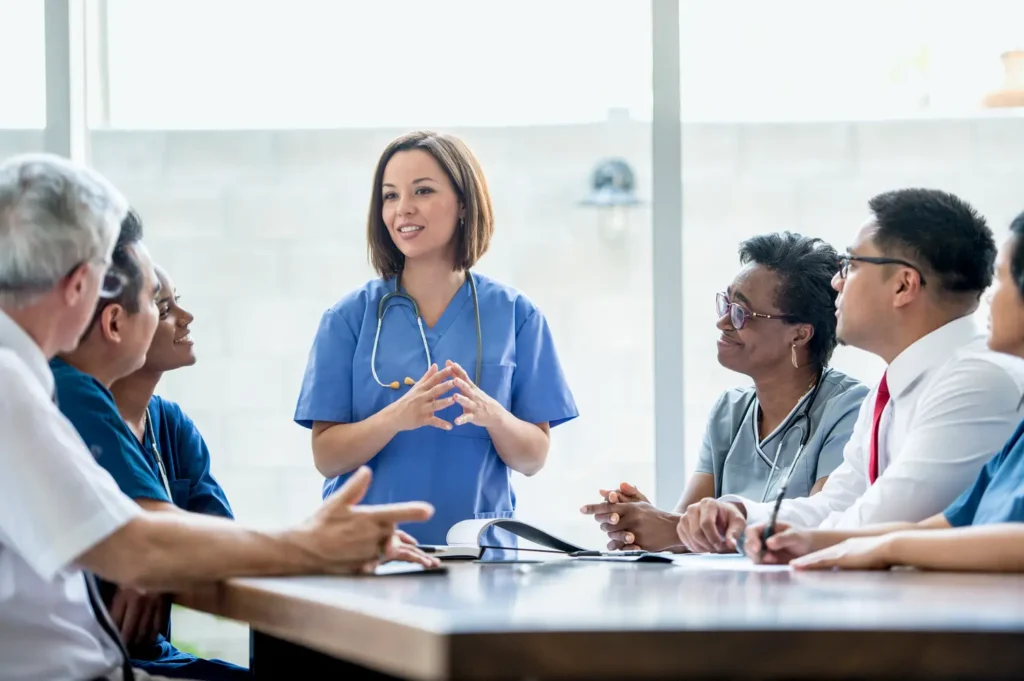A diverse group of healthcare professionals, including a central woman in blue scrubs, attentively listening and interacting during a clinical team meeting in a bright office setting.