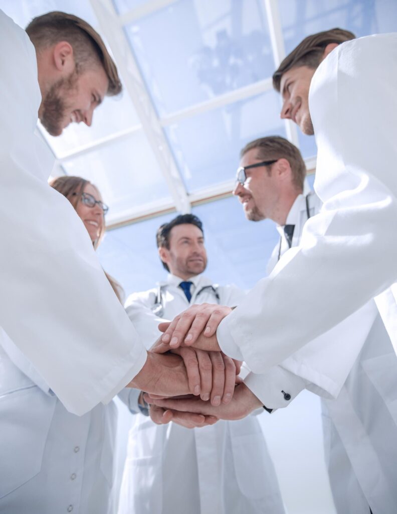 A group of healthcare professionals in white coats stack their hands together in a gesture of teamwork and unity, viewed from a low angle.
