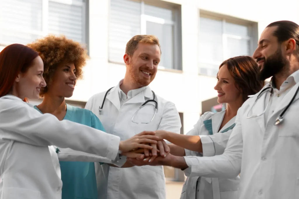 A diverse group of five smiling healthcare professionals, including doctors and nurses, stack their hands together in a gesture of teamwork and unity in a brightly lit clinical environment.
