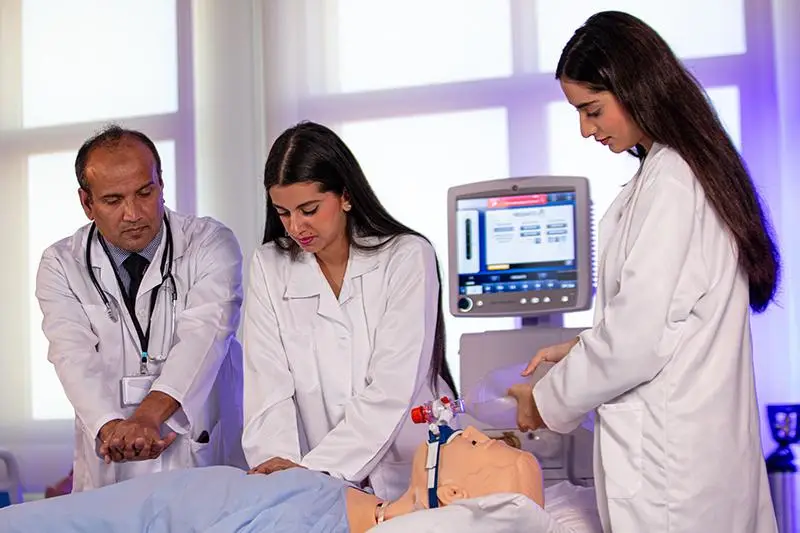 Three healthcare professionals, two women and one man, are attending to a medical training mannequin in a clinical setting, with a monitor displaying vital signs in the background.