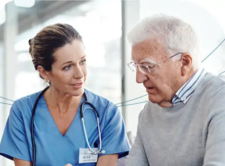 A female nurse in scrubs is attentively listening to an elderly male patient as they discuss his health in a bright primary care office. The nurse is holding a digital tablet.