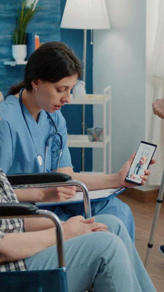 A female nurse in blue scrubs shows a tablet to an elderly male patient in a wheelchair, displaying another doctor on the screen, inside a home setting for advanced primary care.