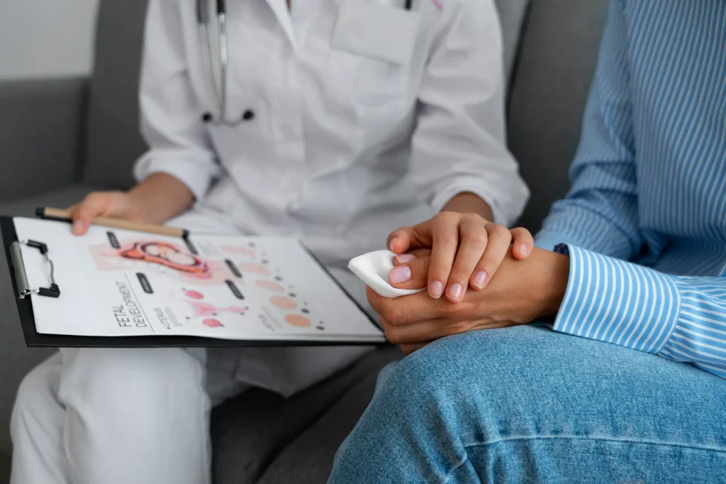 A healthcare professional holding a patient's hand, offering comfort during a clinical consultation, with a clipboard displaying a female reproductive system diagram.