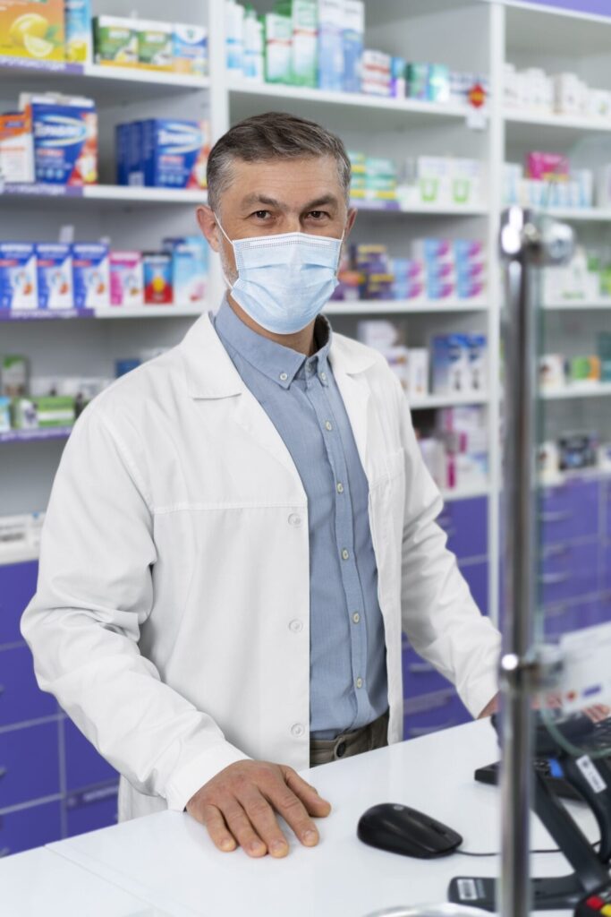 A mature male pharmacist wearing a face mask and a white coat stands behind the checkout counter in a clinical pharmacy, surrounded by shelves filled with medical products.