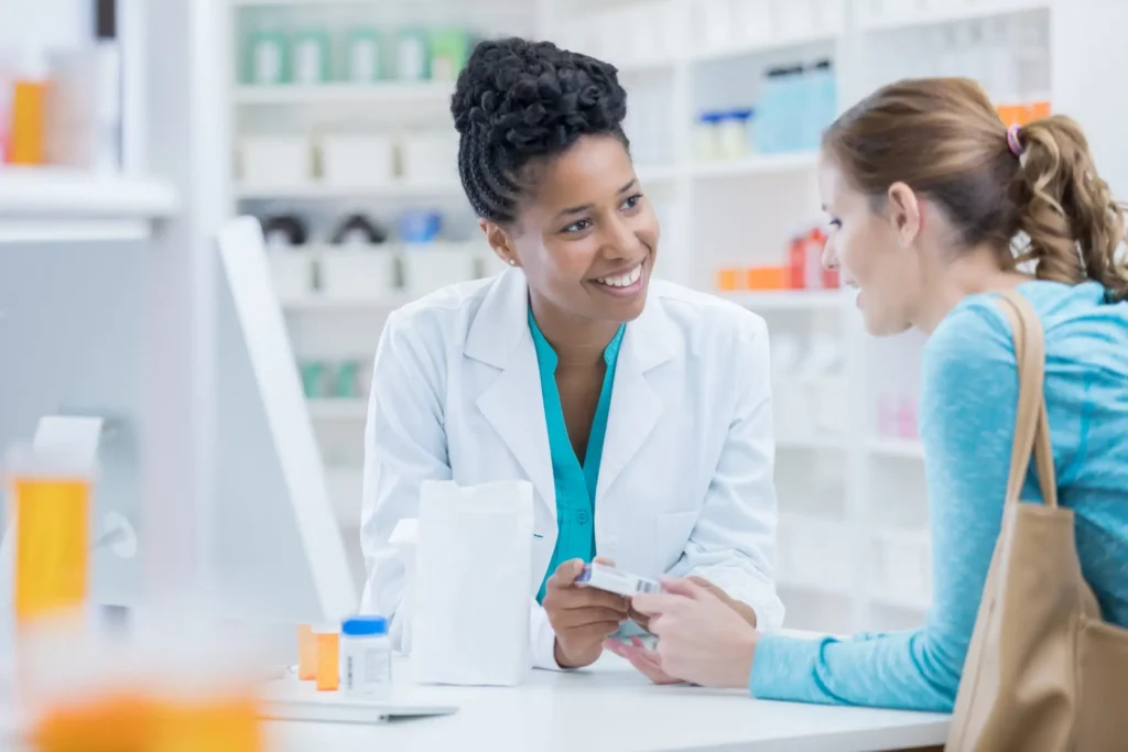 A pharmacist, a Black woman with a friendly smile wearing a white coat, counseling a female patient over a counter in a brightly lit Advanced Primary Care pharmacy.