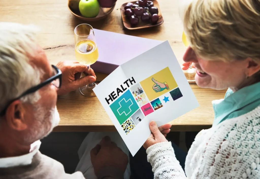 Two senior adults looking at a brochure labeled "Advanced Primary Care" with colorful icons related to healthcare, sitting at a wooden table with fruit and wine.