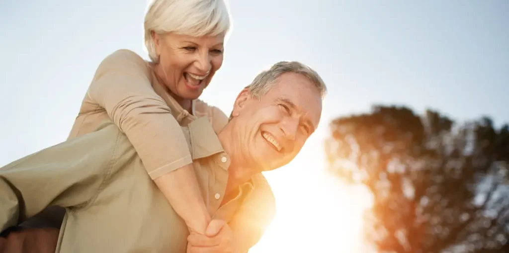 An elderly man giving a piggyback ride to an elderly woman, both laughing joyfully with a softly lit, sunlit backdrop in an Advanced Primary Care setting.