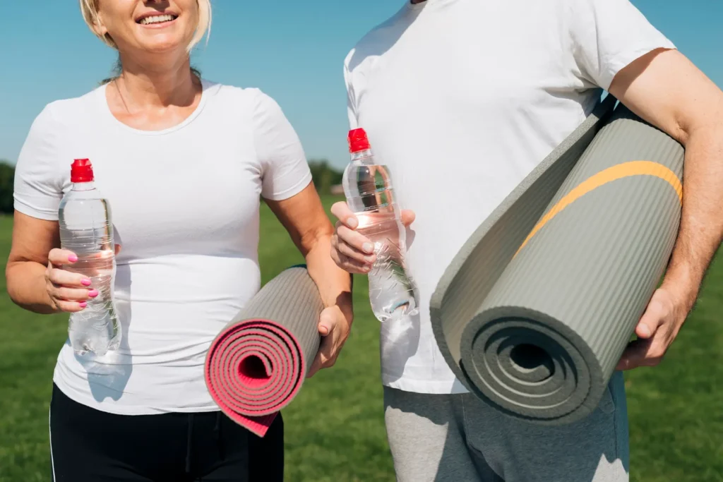 Two senior adults holding water bottles and yoga mats, standing in a sunny park, dressed in sportswear, smiling and ready for their Advanced Primary Care workout session.