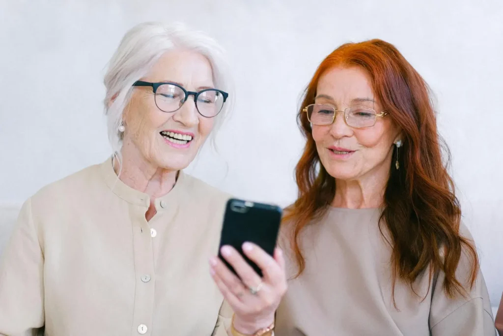 Two elderly women, one with white hair and the other with red hair, smiling and looking at a smartphone together in a brightly lit room, discussing Advanced Primary Care options.