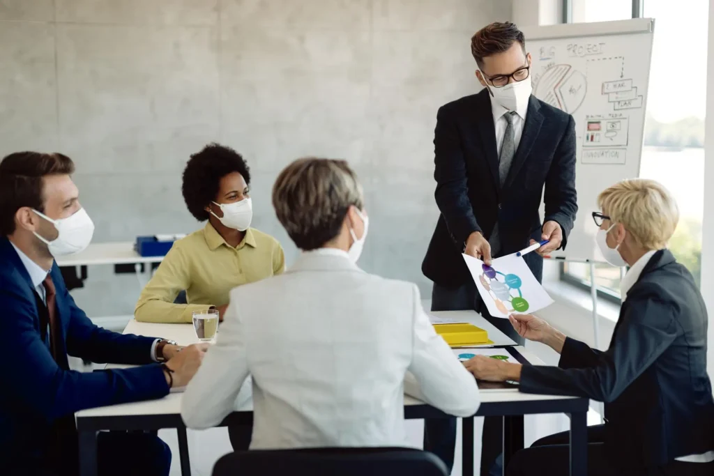 Businessman discussing charts with coworkers in a masked meeting room during the COVID-19 pandemic.