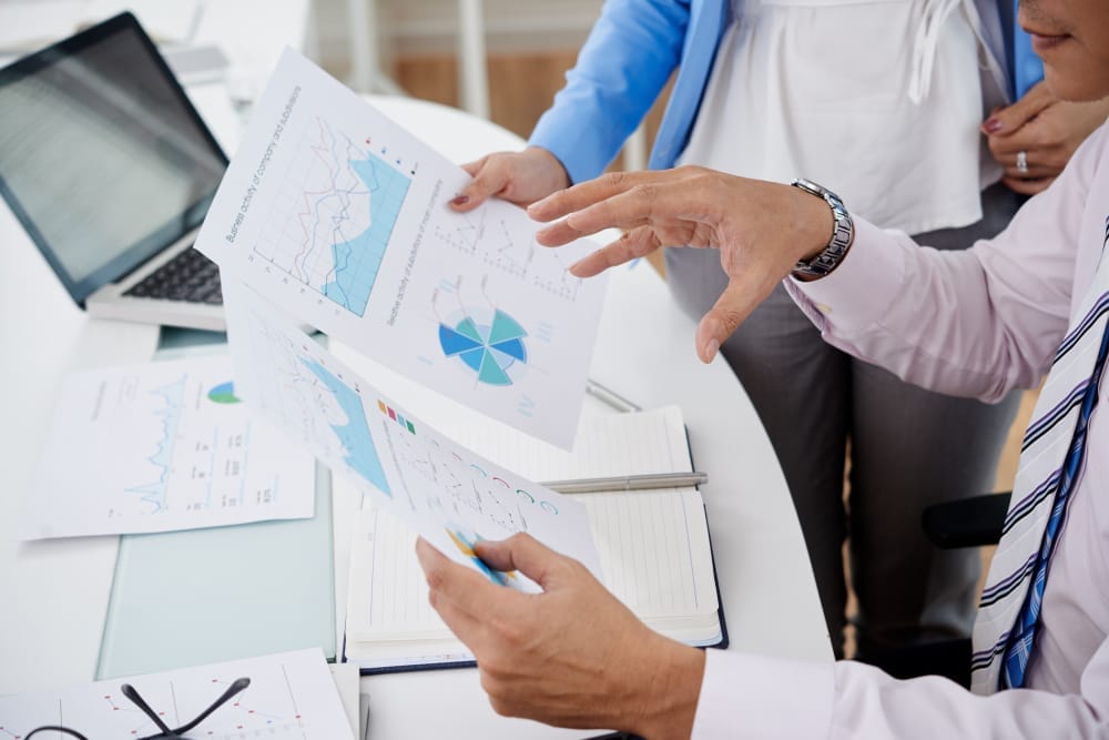 Colleagues discussing charts and data from clinical trials during a business meeting, close-up of hands pointing at documents, with a laptop open in the background.