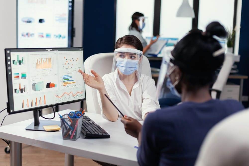 Two healthcare workers, one wearing a face shield and mask, discussing data on a computer monitor in a modern office setting.
