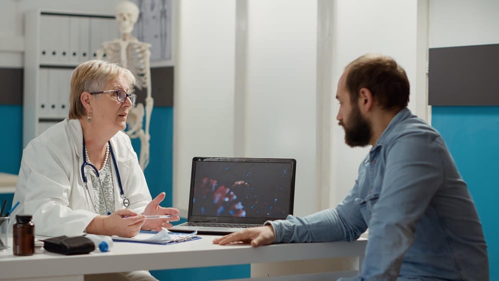 A female doctor in a white coat talks to a male participant in a clinic office with a laptop open between them and a skeleton model in the background.