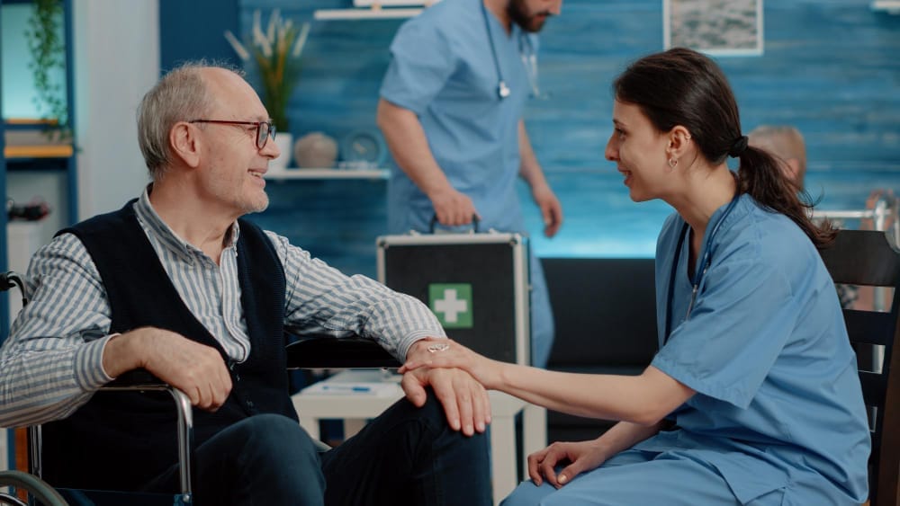 A senior man in a wheelchair and a female nurse sit facing each other, smiling and holding hands in a setting of clinical trials, with a caretaker adjusting a second wheelchair in the background.